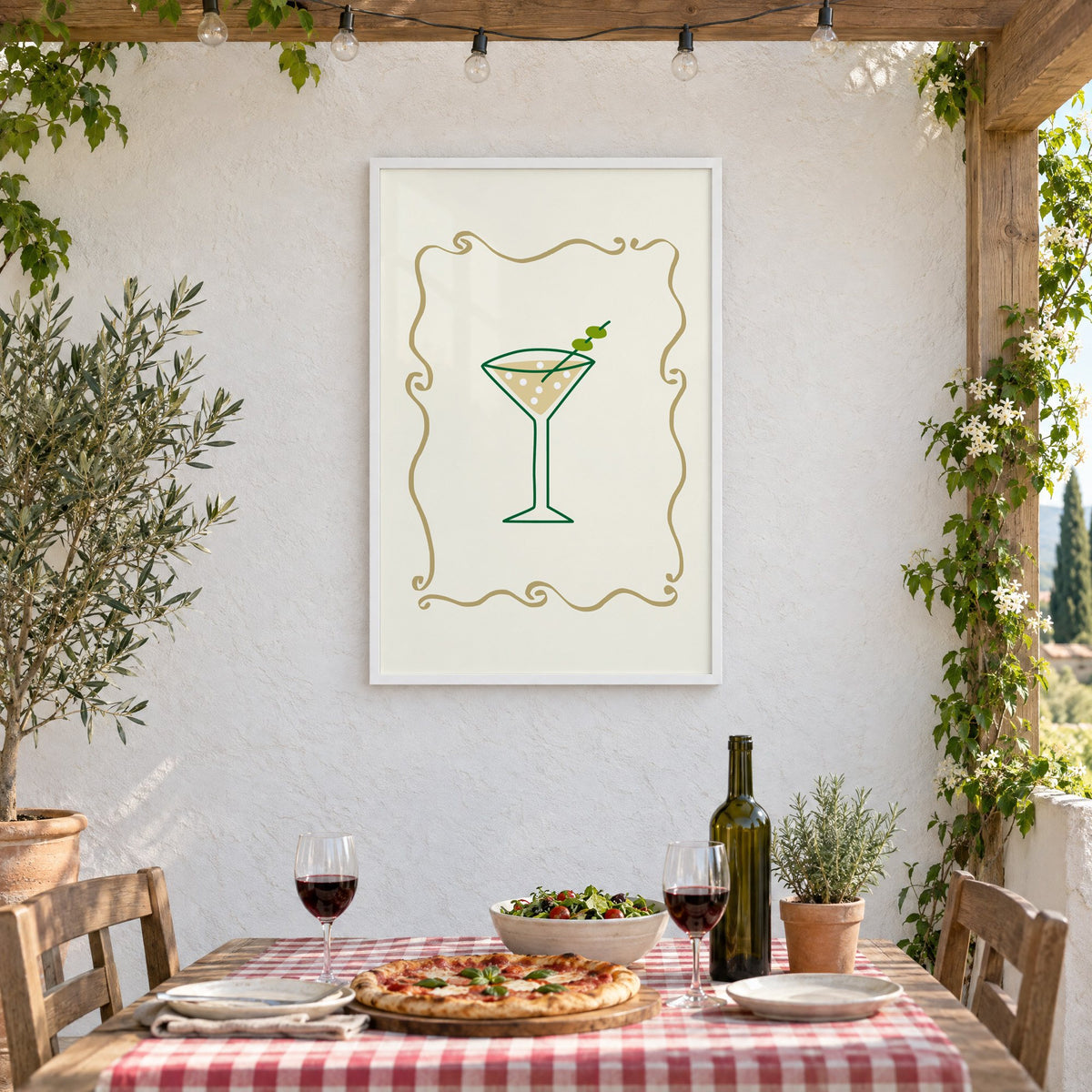 Olive martini in gold scroll frame print in a white frame on a stucco wall above a checked-cloth table with pizza and red wine