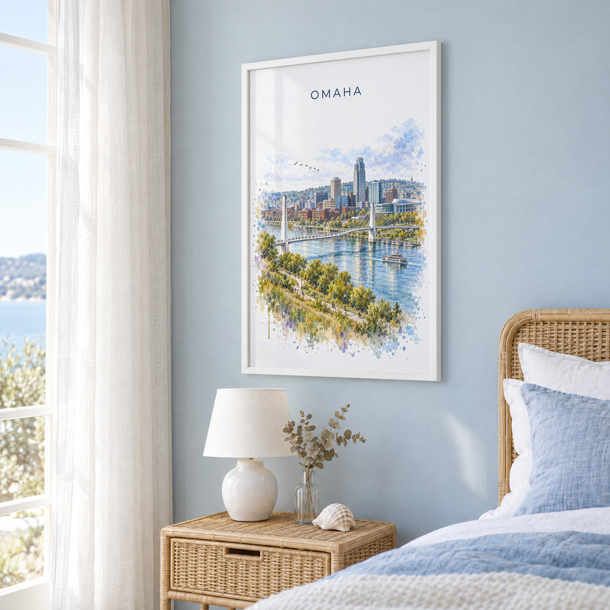 Omaha watercolor print in a white frame on a pale blue wall beside a rattan bed in a coastal bedroom