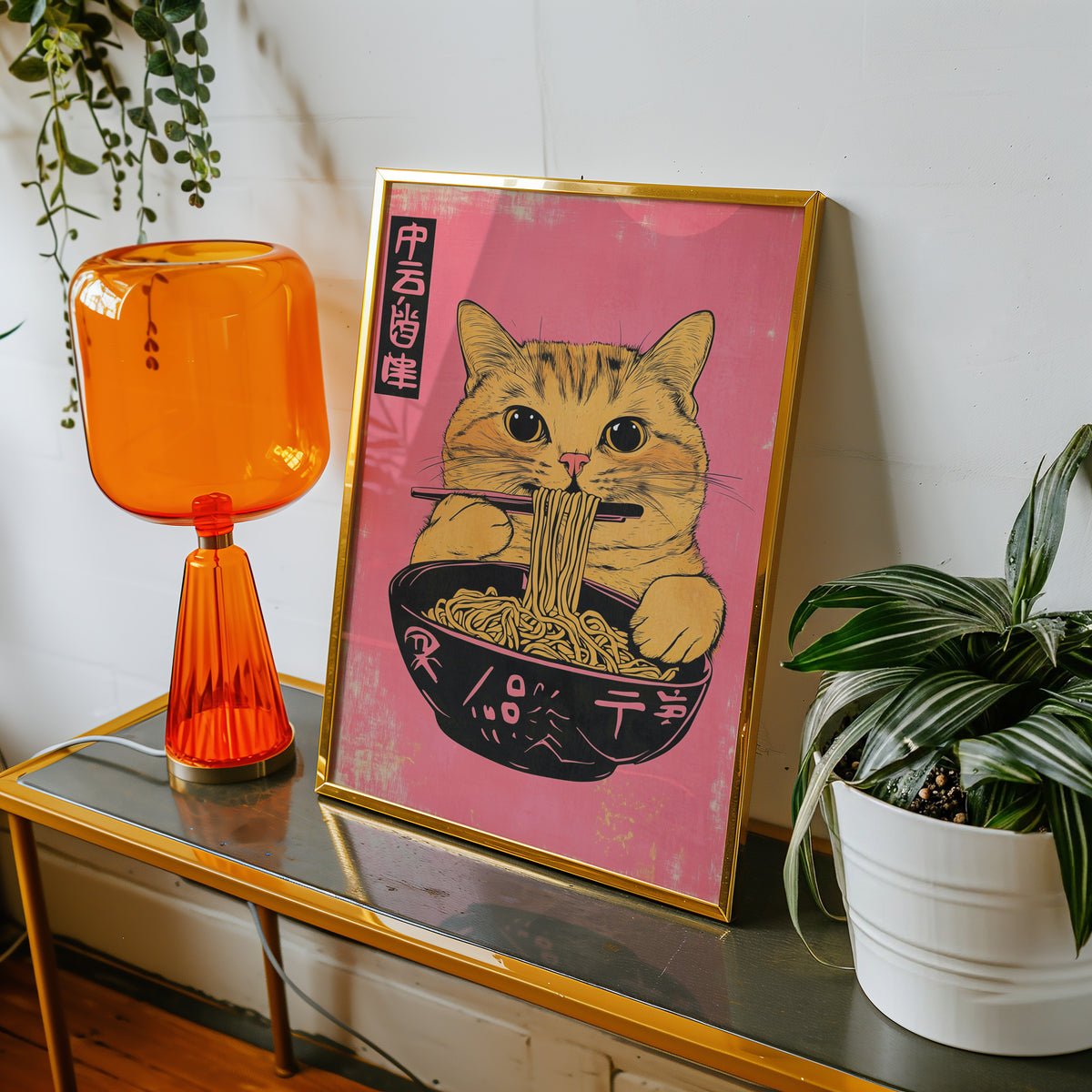 Orange tabby cat slurping ramen print shown in a gold frame on a console beside an orange glass lamp