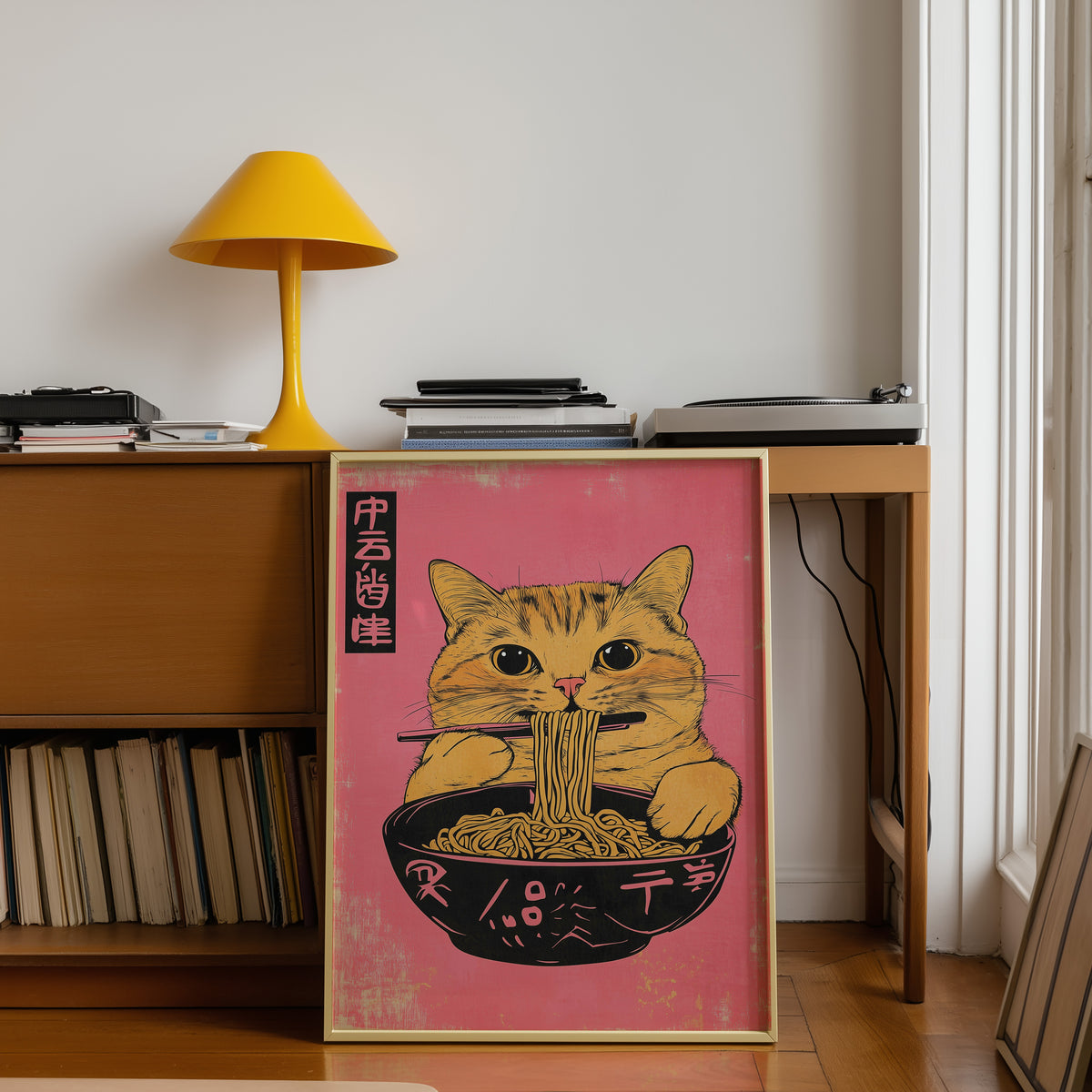 Orange tabby cat slurping ramen print shown in a gold frame below a teak desk with a yellow lamp