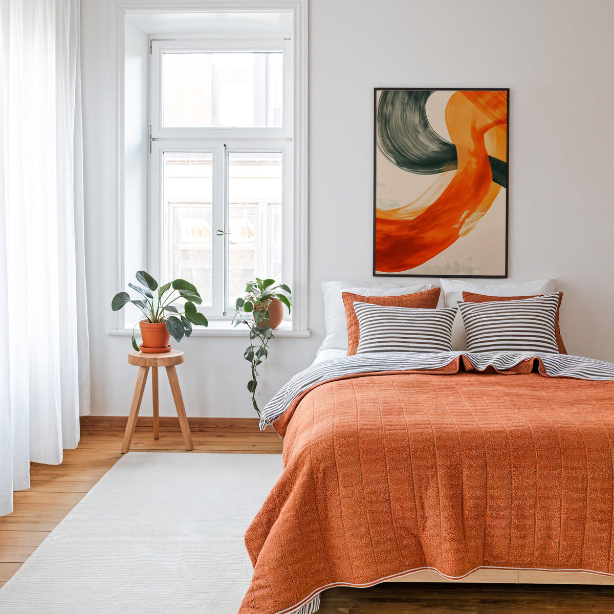 Orange and charcoal brushstroke abstract print in a thin black frame above a bed with a rust orange quilt