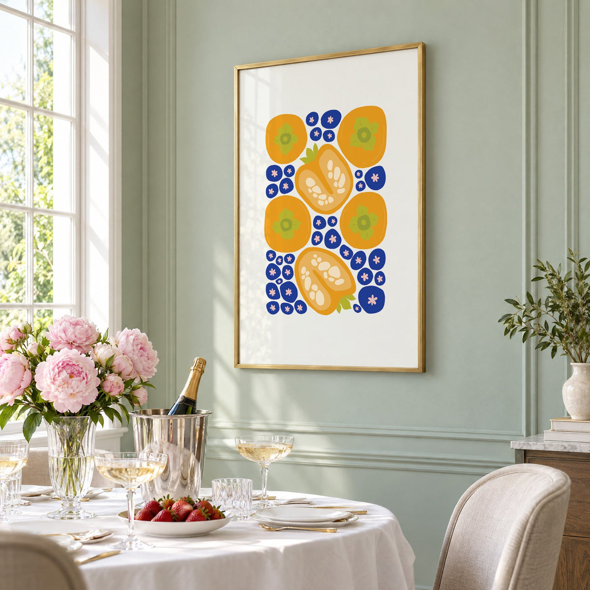 orange persimmons cobalt blueberries pattern print in a brass frame on a sage wall above a brunch table with champagne and peonies, seen at an angle