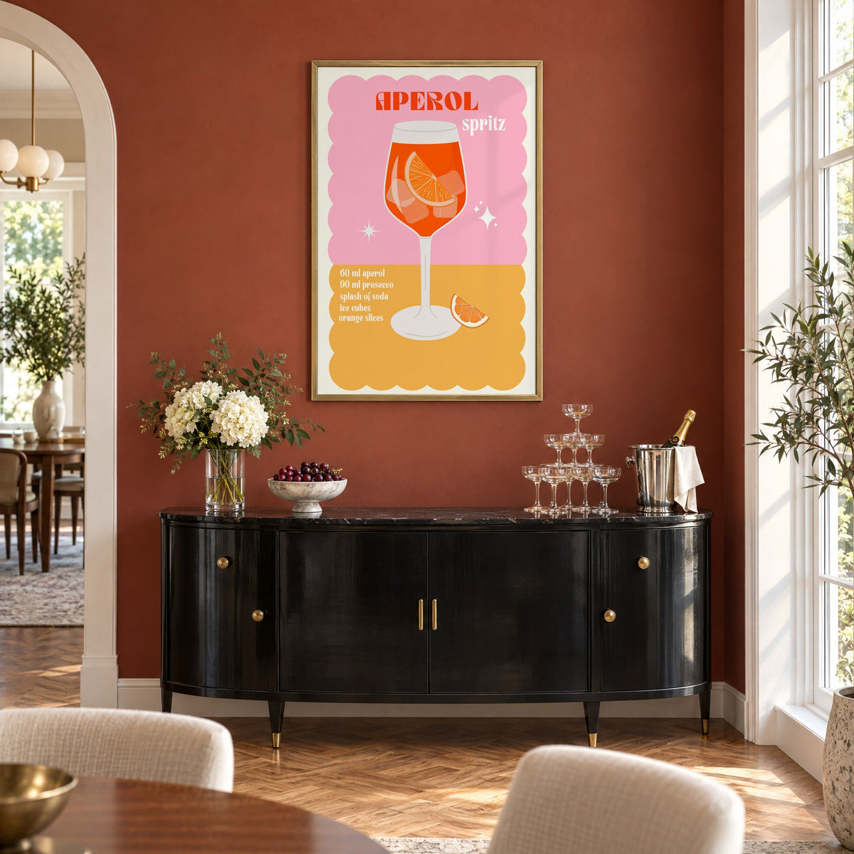 Orange spritz scalloped pink print in a brass frame on a terracotta wall above a black buffet with a champagne tower