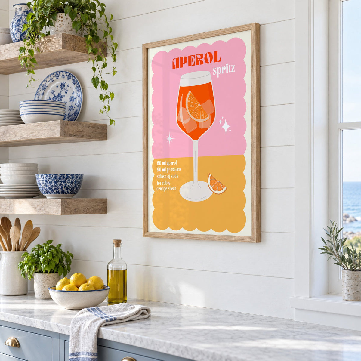 Orange spritz scalloped pink print in an oak frame in a white coastal kitchen beside blue and white plates