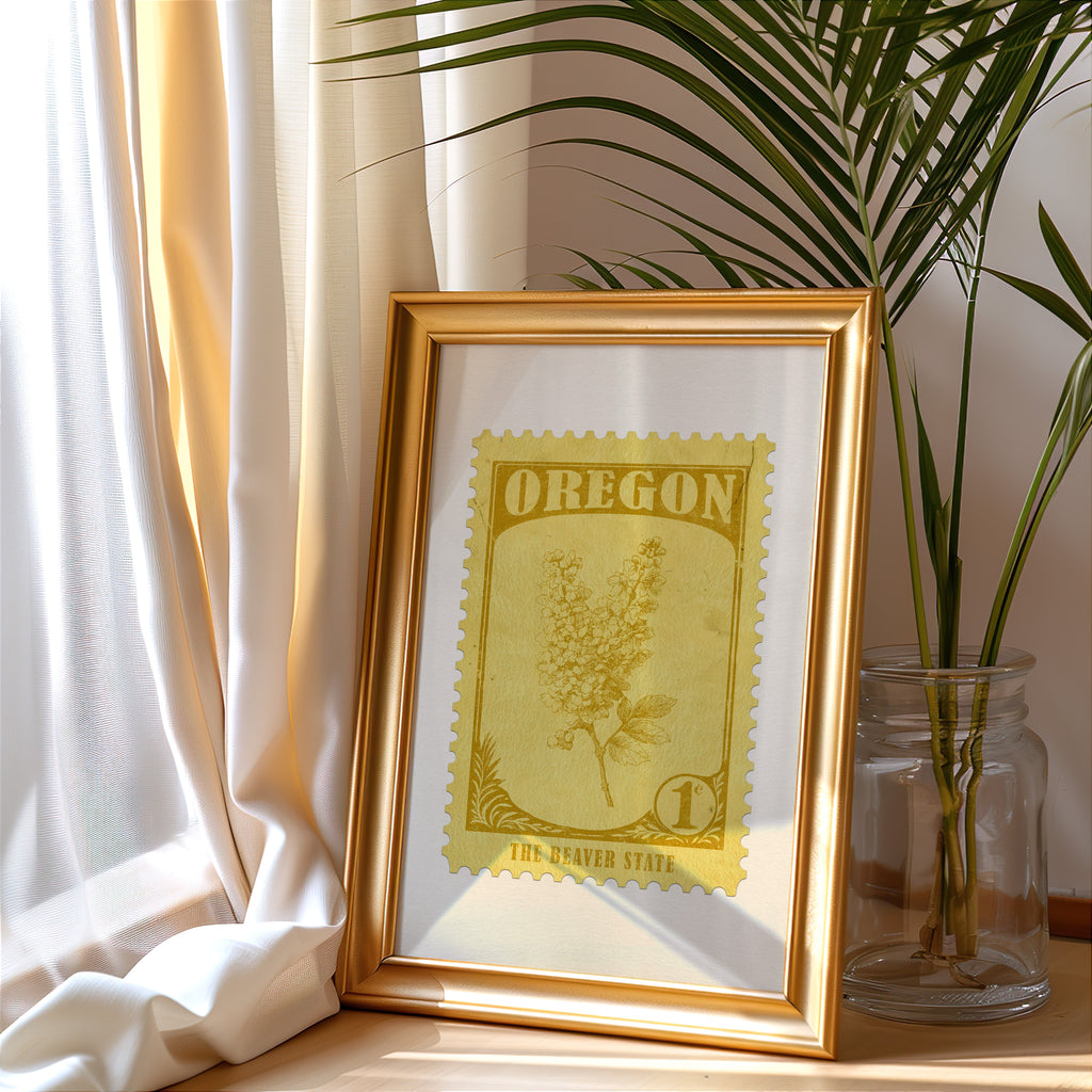 Oregon stamp print in a gold frame leaning by a cream curtain with a palm in a glass jar