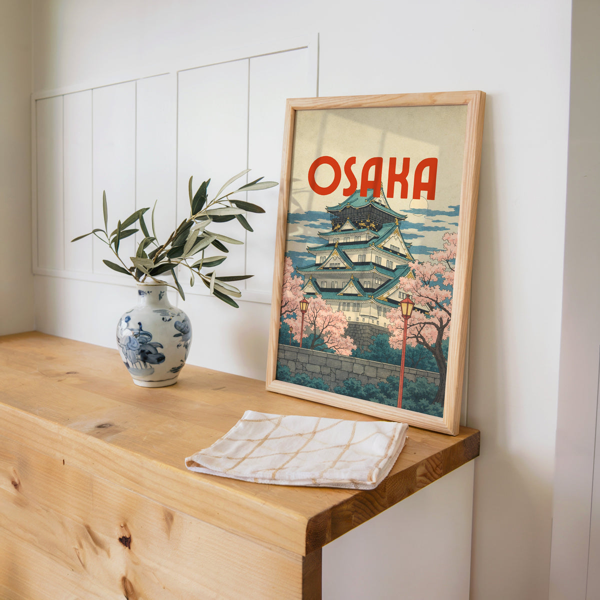 Osaka Castle with teal roofs print in a pine frame on a wooden counter beside a blue and white porcelain vase