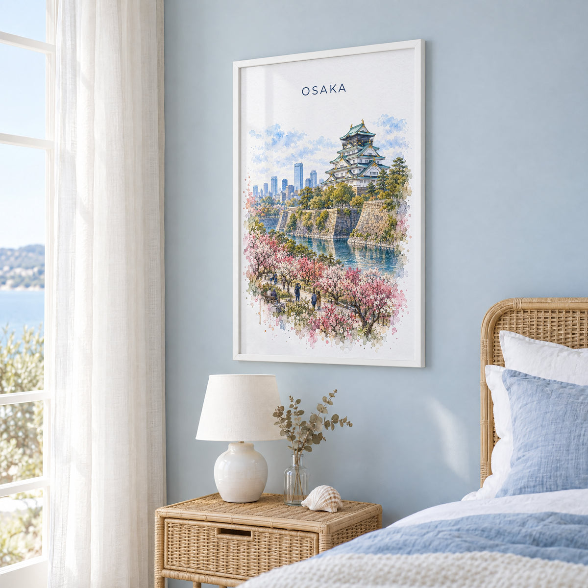 Osaka watercolor print in a white frame on a pale blue wall beside a rattan bed in a coastal bedroom