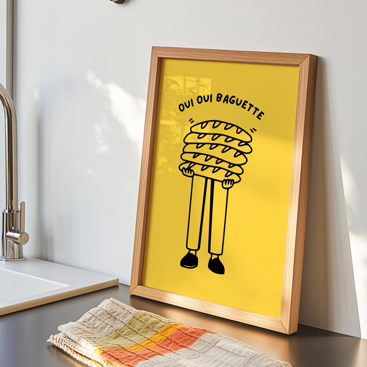 Oui Oui Baguette print in an oak frame by the kitchen sink with a striped tea towel