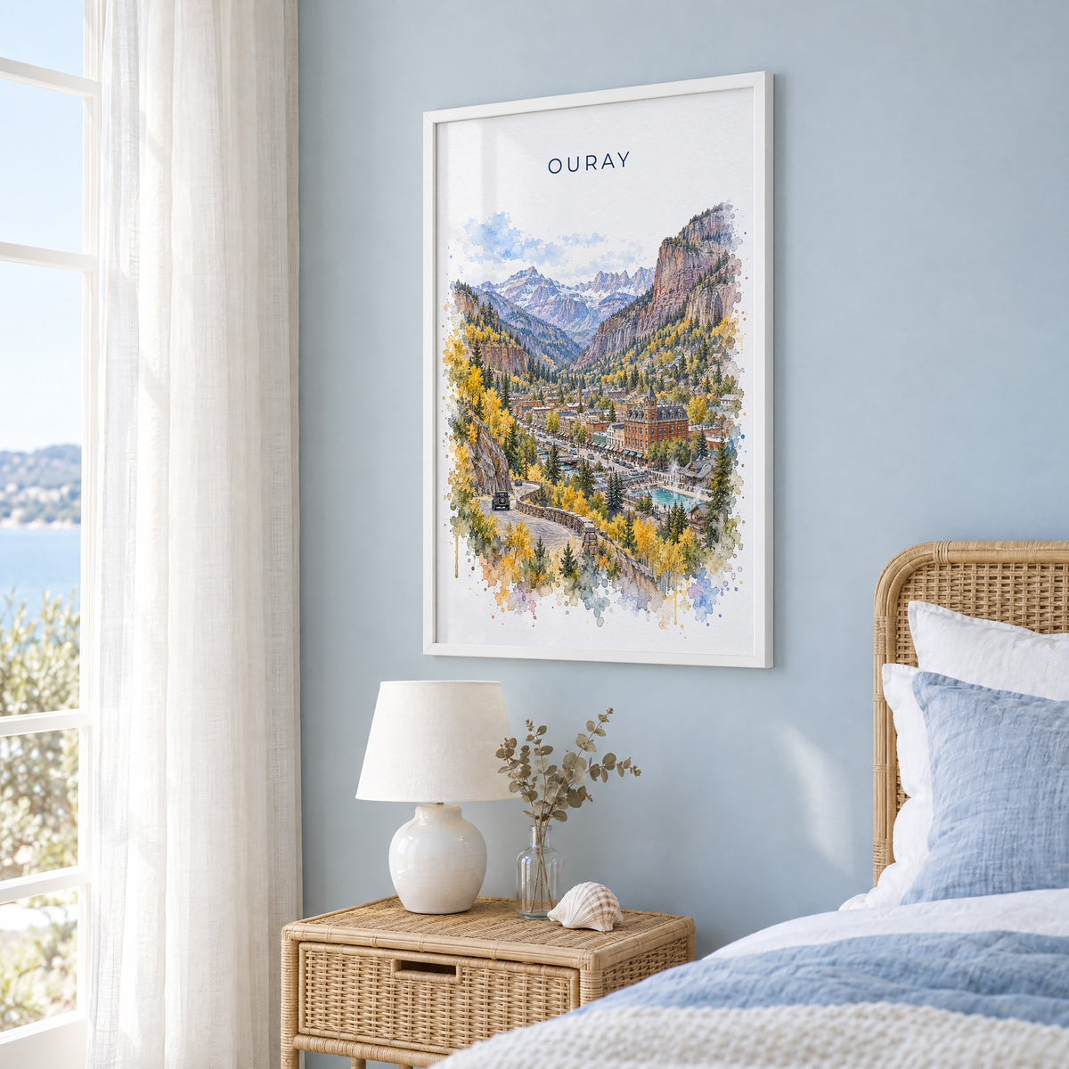 Ouray watercolor print in a white frame on a pale blue wall beside a rattan bed in a coastal bedroom