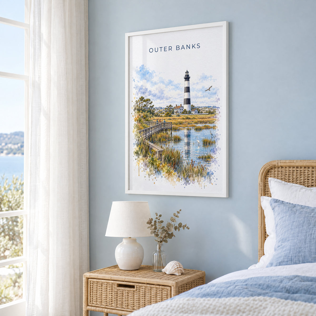 Outer Banks watercolor print in a white frame on a pale blue wall beside a rattan bed in a coastal bedroom