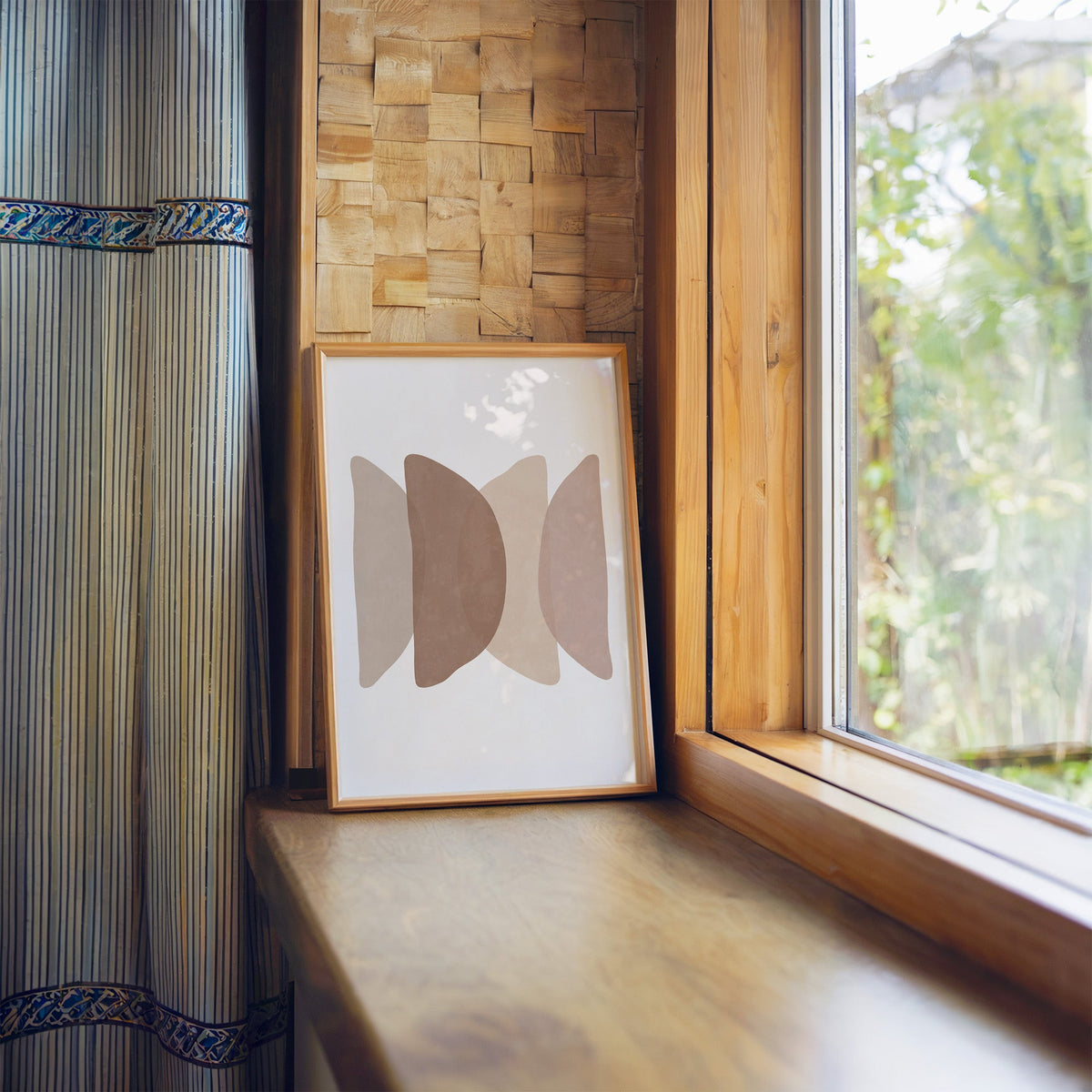 Overlapping blush pebble shapes boho print in a light wood frame leaning on a wooden window sill by a garden view