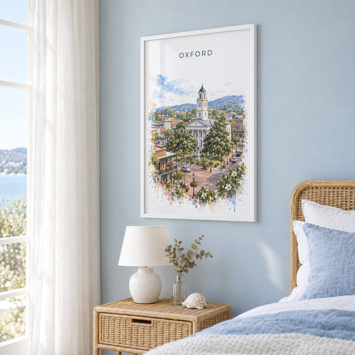 Oxford, Mississippi watercolor print in a white frame on a pale blue wall beside a rattan bed in a coastal bedroom