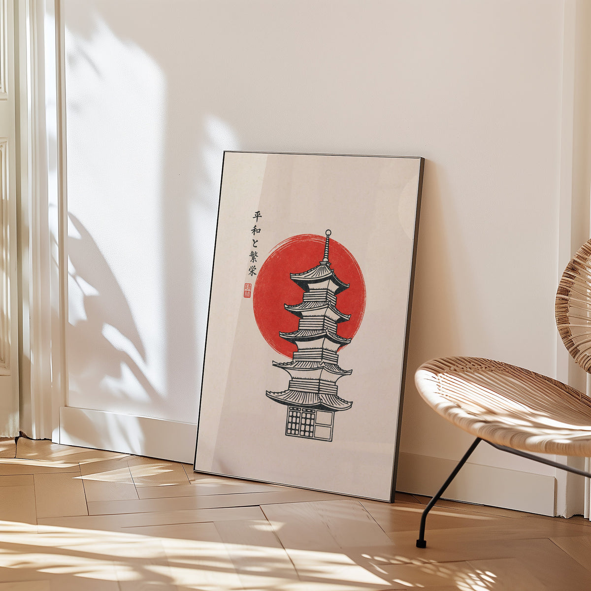 pagoda line art with red sun print in a black frame leaning on a herringbone floor beside a woven rattan chair