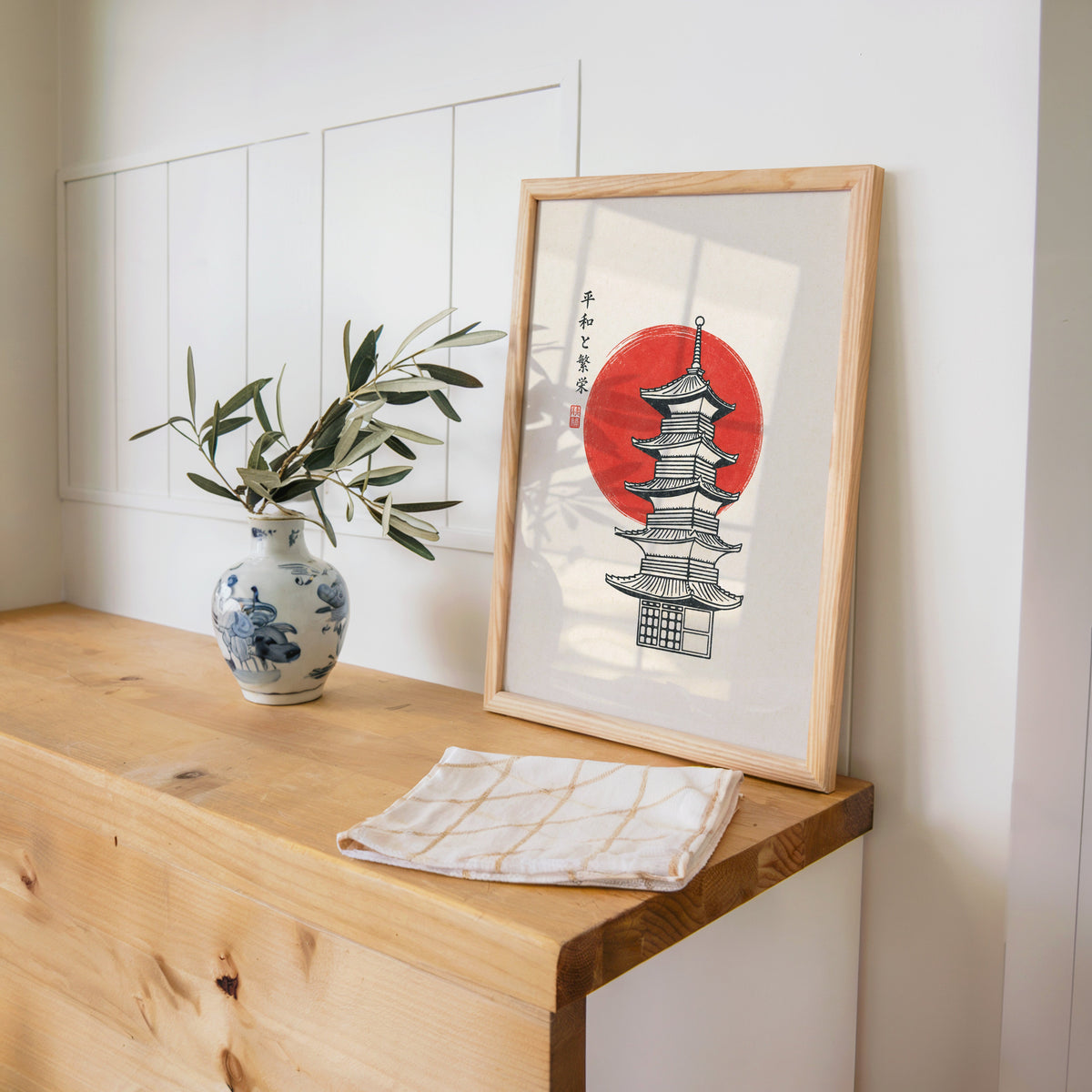 pagoda line art with red sun print in a pine frame on a wooden counter beside a blue and white porcelain vase