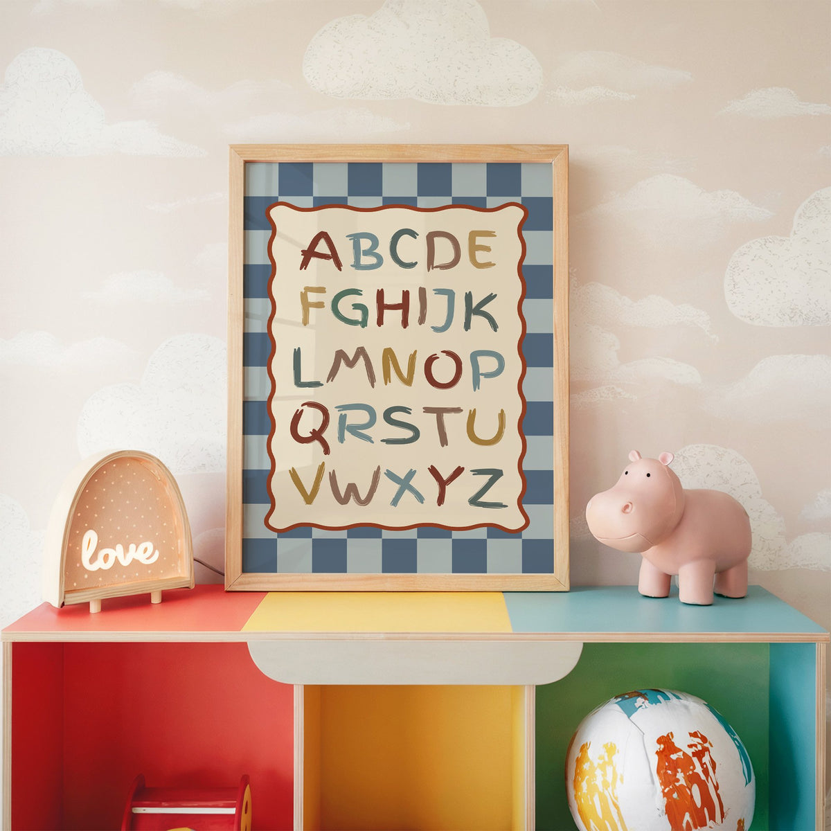 Painted alphabet wavy frame nursery print in an oak frame on a rainbow toy shelf against cloud wallpaper