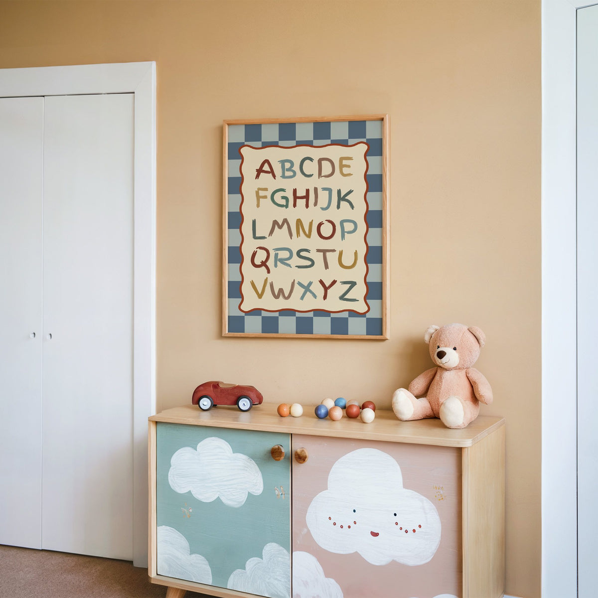 Painted alphabet wavy frame nursery print in an oak frame on a peach wall above a cloud-painted cabinet