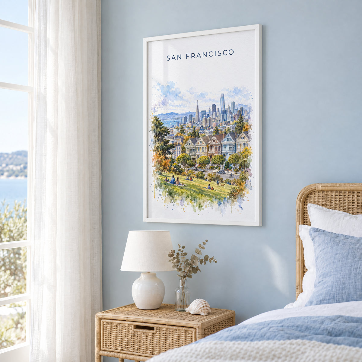 Painted Ladies San Francisco watercolor print in a white frame on a pale blue wall beside a rattan bed in a coastal bedroom