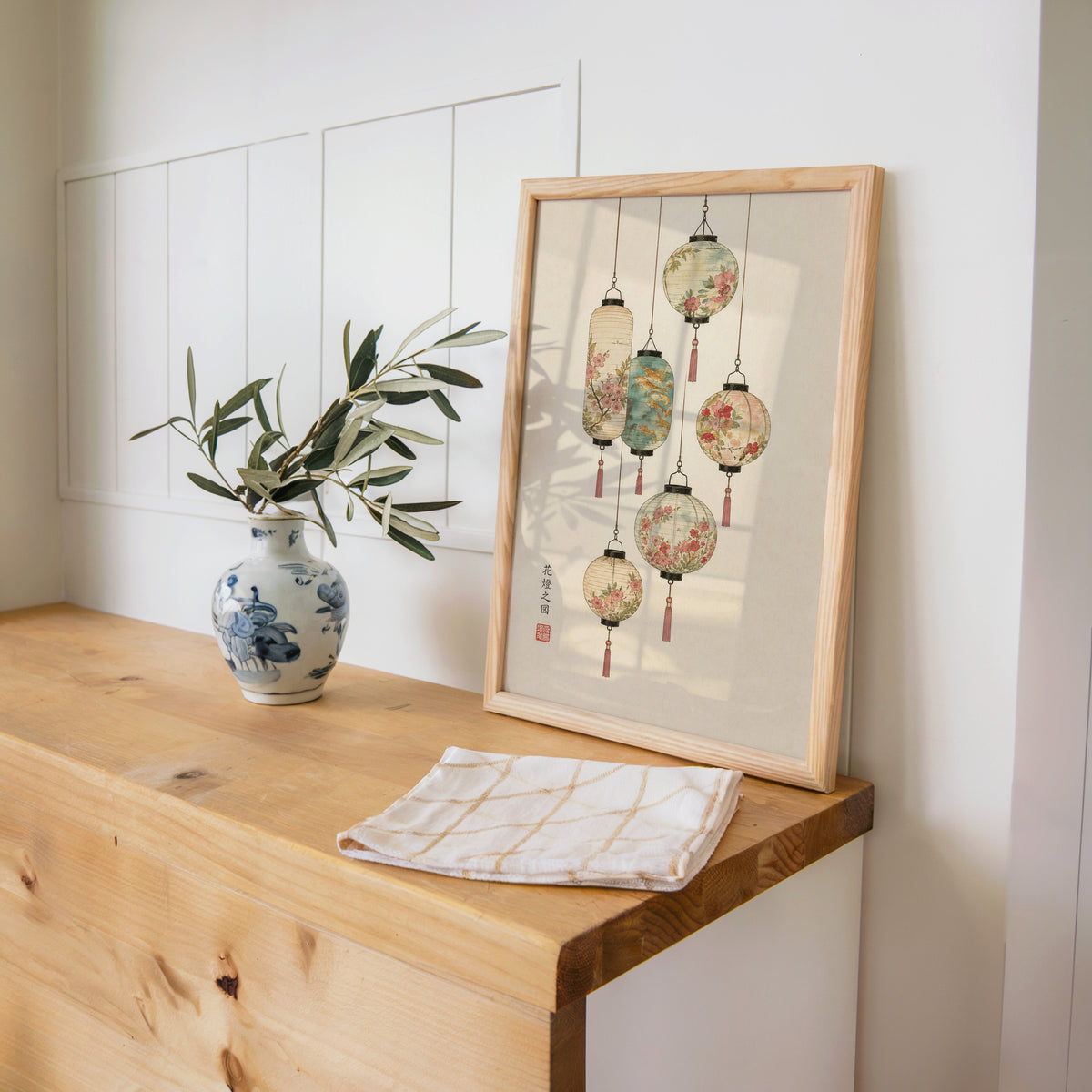 painted paper lanterns with flowers print in a pine frame on a wooden counter beside a blue and white porcelain vase