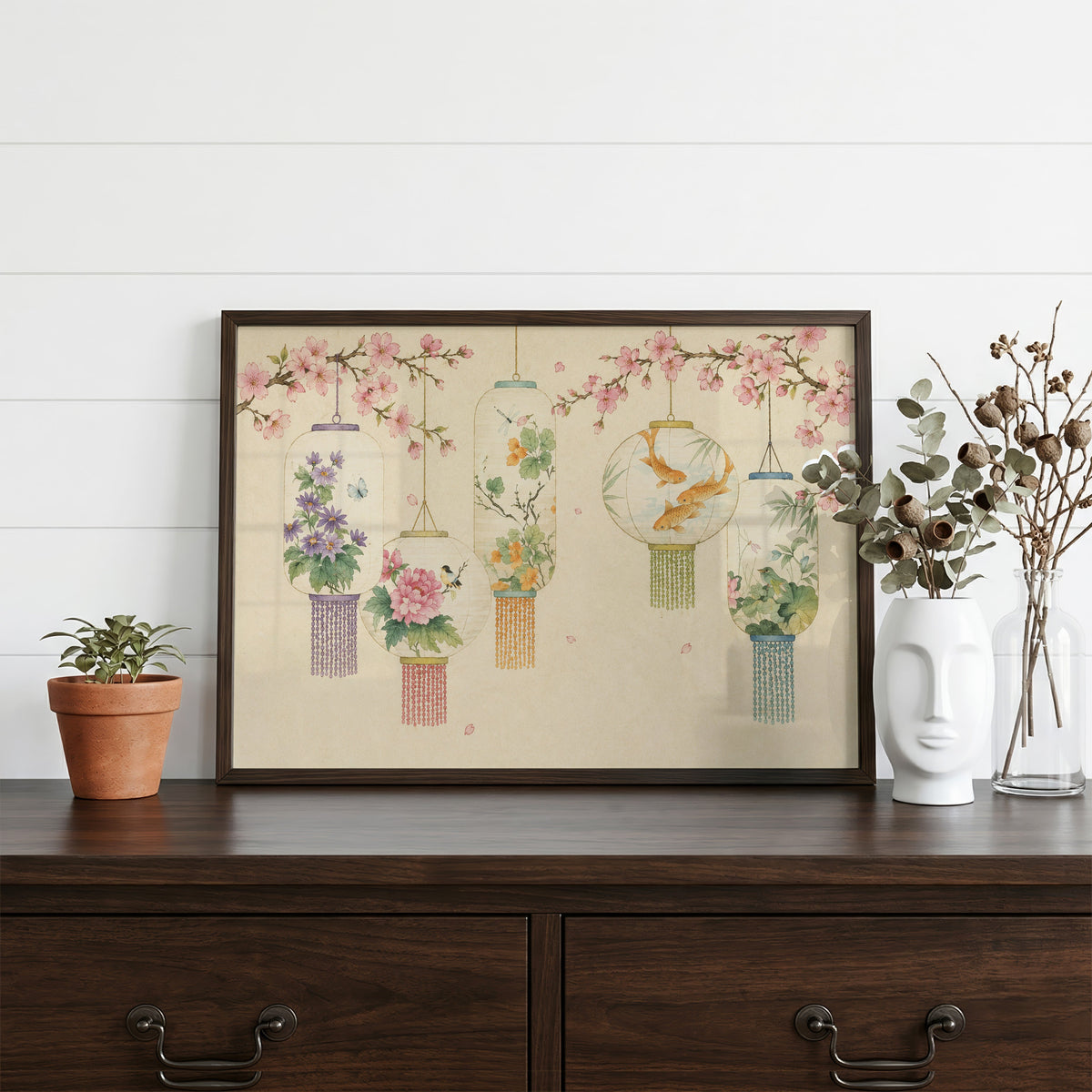 Five painted lanterns under cherry blossom print in a dark walnut frame on a dark wood dresser beside a white face vase