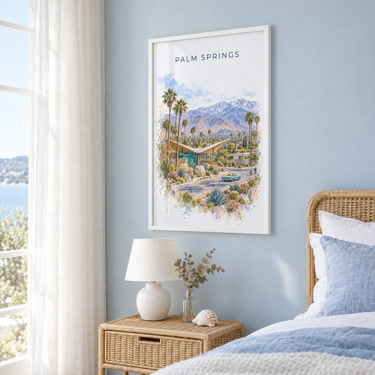 Palm Springs watercolor print in a white frame on a pale blue wall beside a rattan bed in a coastal bedroom