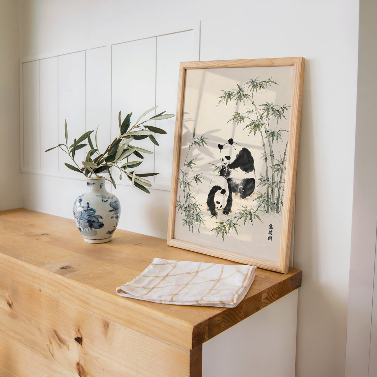 two pandas with bamboo ink painting print in a pine frame on a wooden counter beside a blue and white porcelain vase