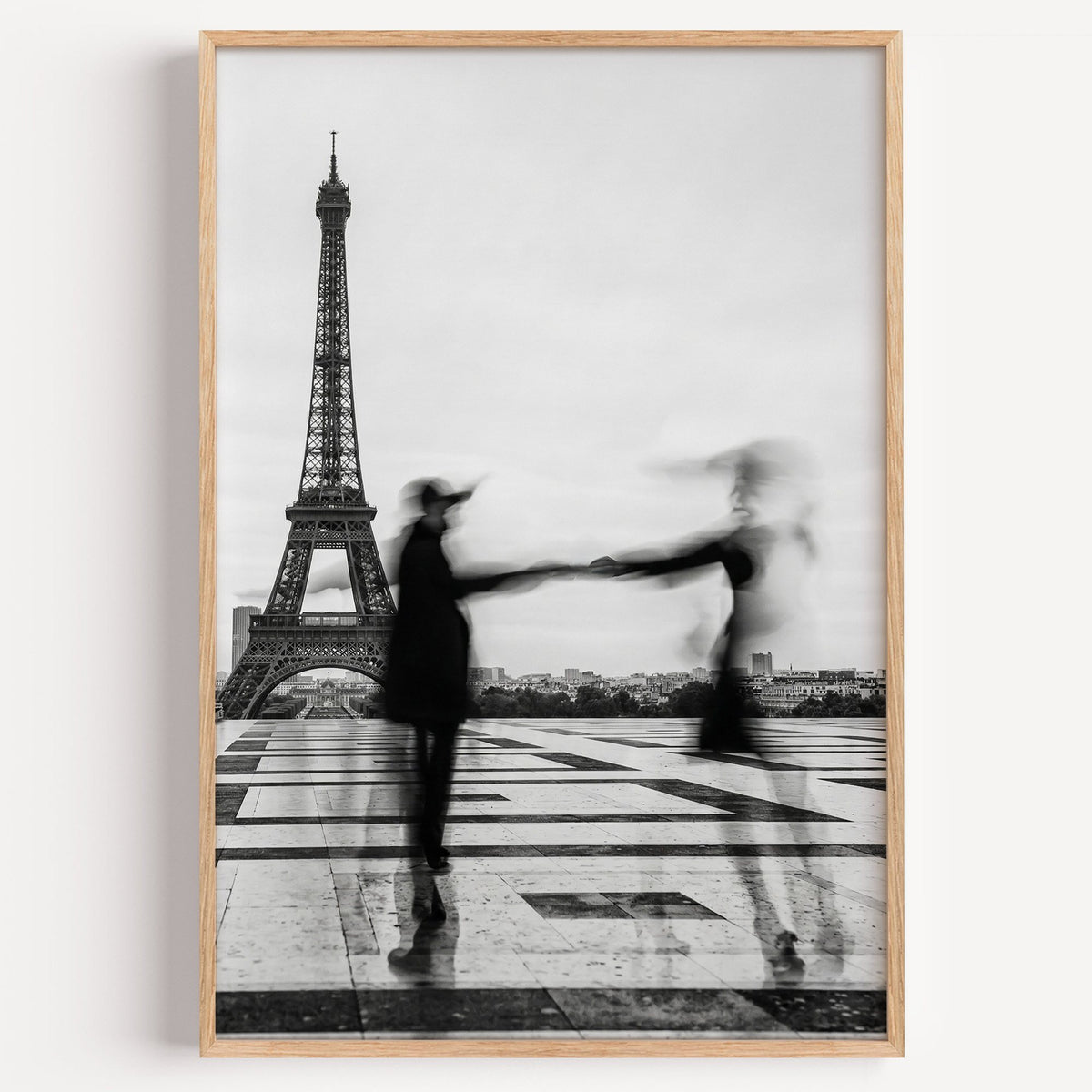 Blurred couple twirling near the Eiffel Tower print in a light oak frame on a plain white wall