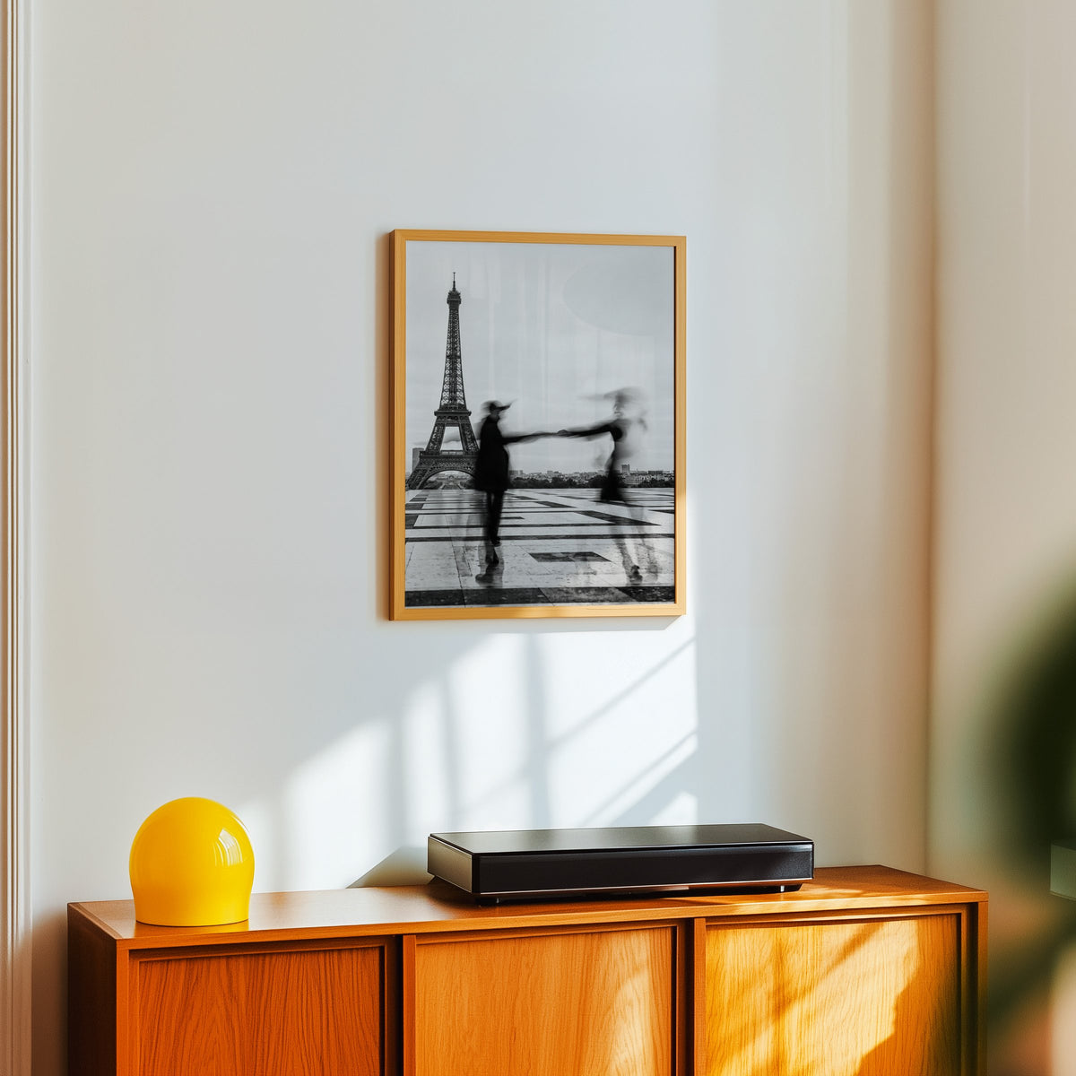 Blurred couple twirling near the Eiffel Tower print in an oak frame above a teak sideboard with a yellow glass lamp
