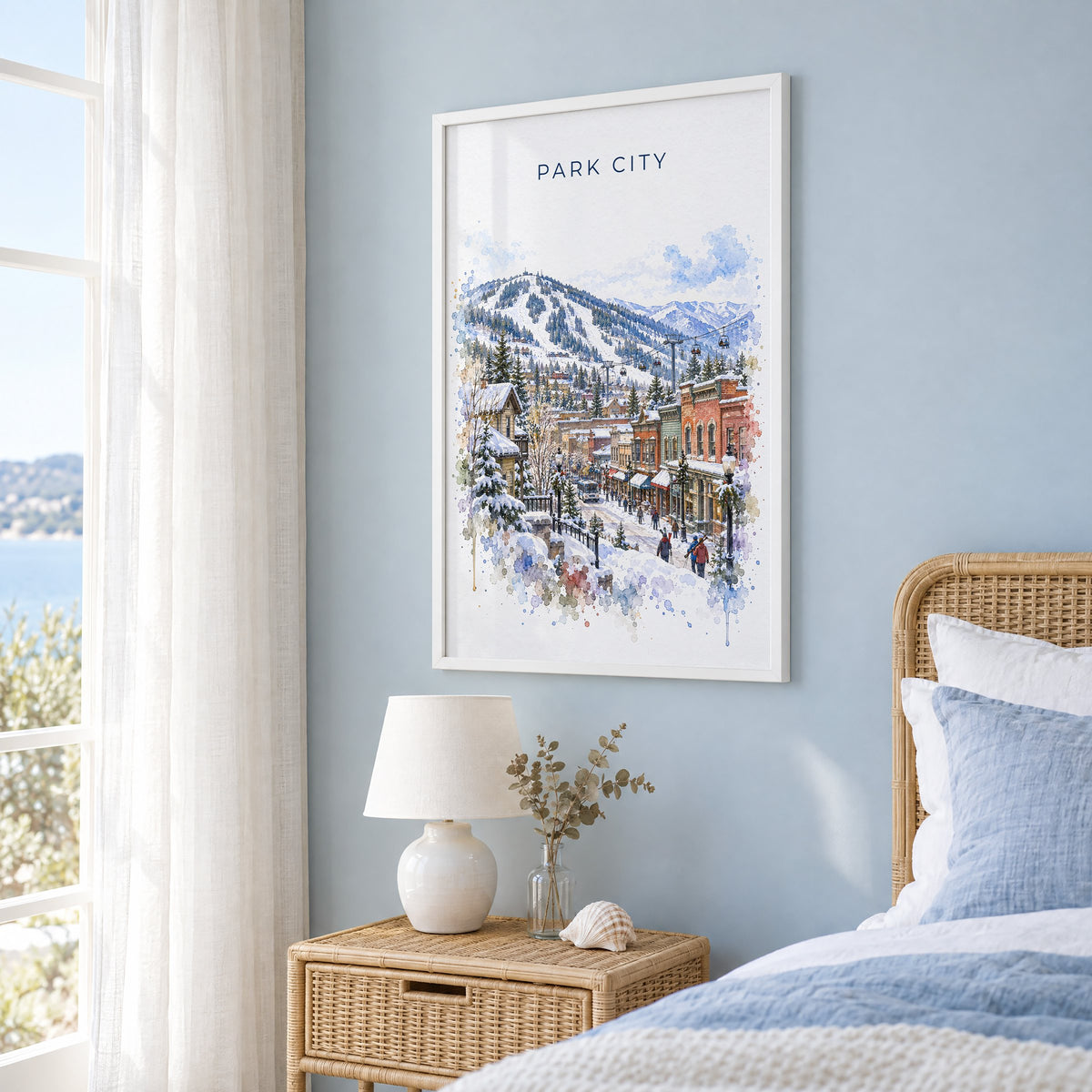 Park City watercolor print in a white frame on a pale blue wall beside a rattan bed in a coastal bedroom