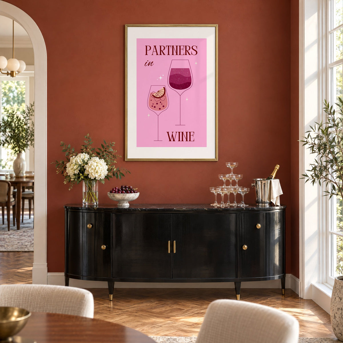 Partners in Wine two glasses pink print in a brass frame on a terracotta wall above a black buffet with a champagne tower