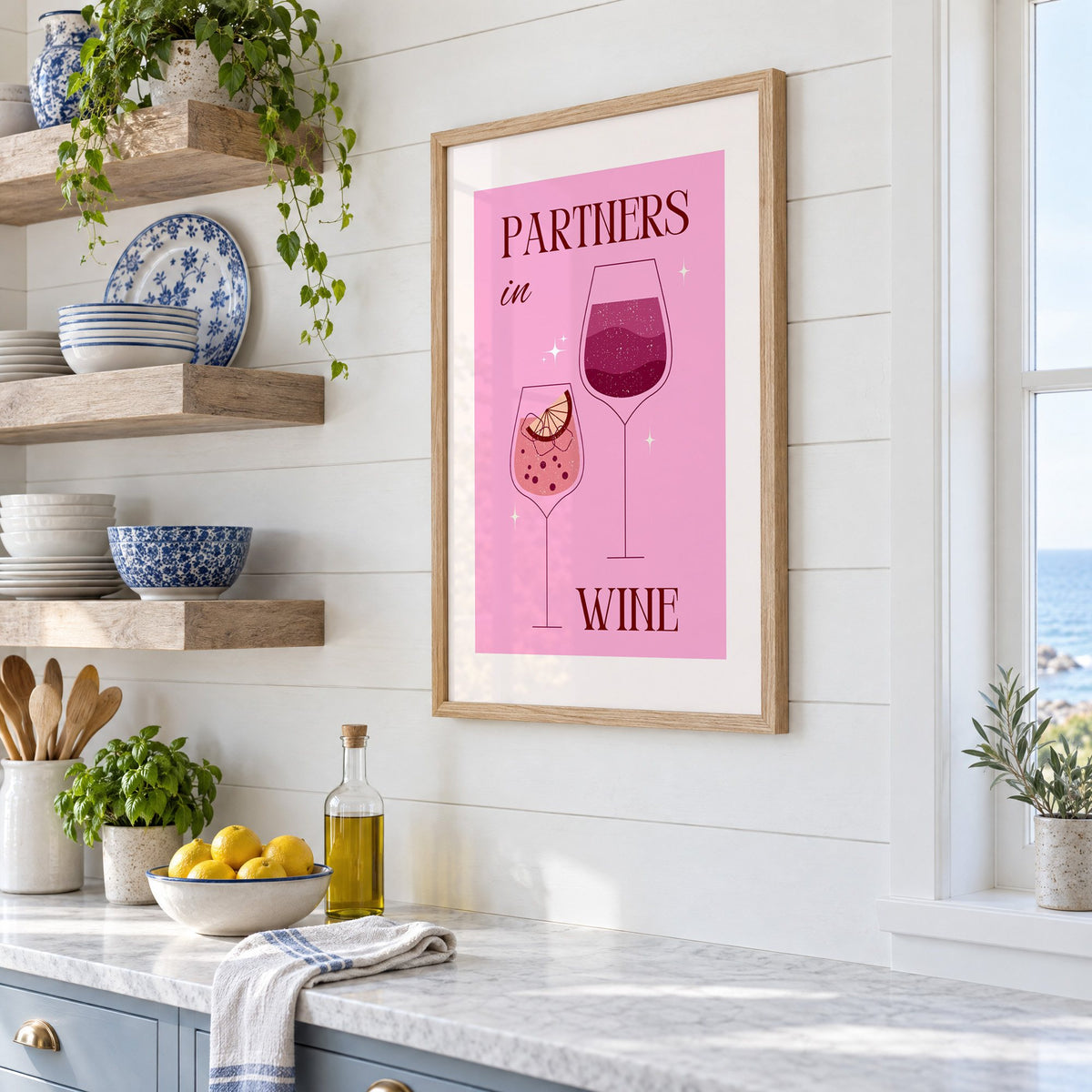 Partners in Wine two glasses pink print in an oak frame in a white coastal kitchen beside blue and white plates
