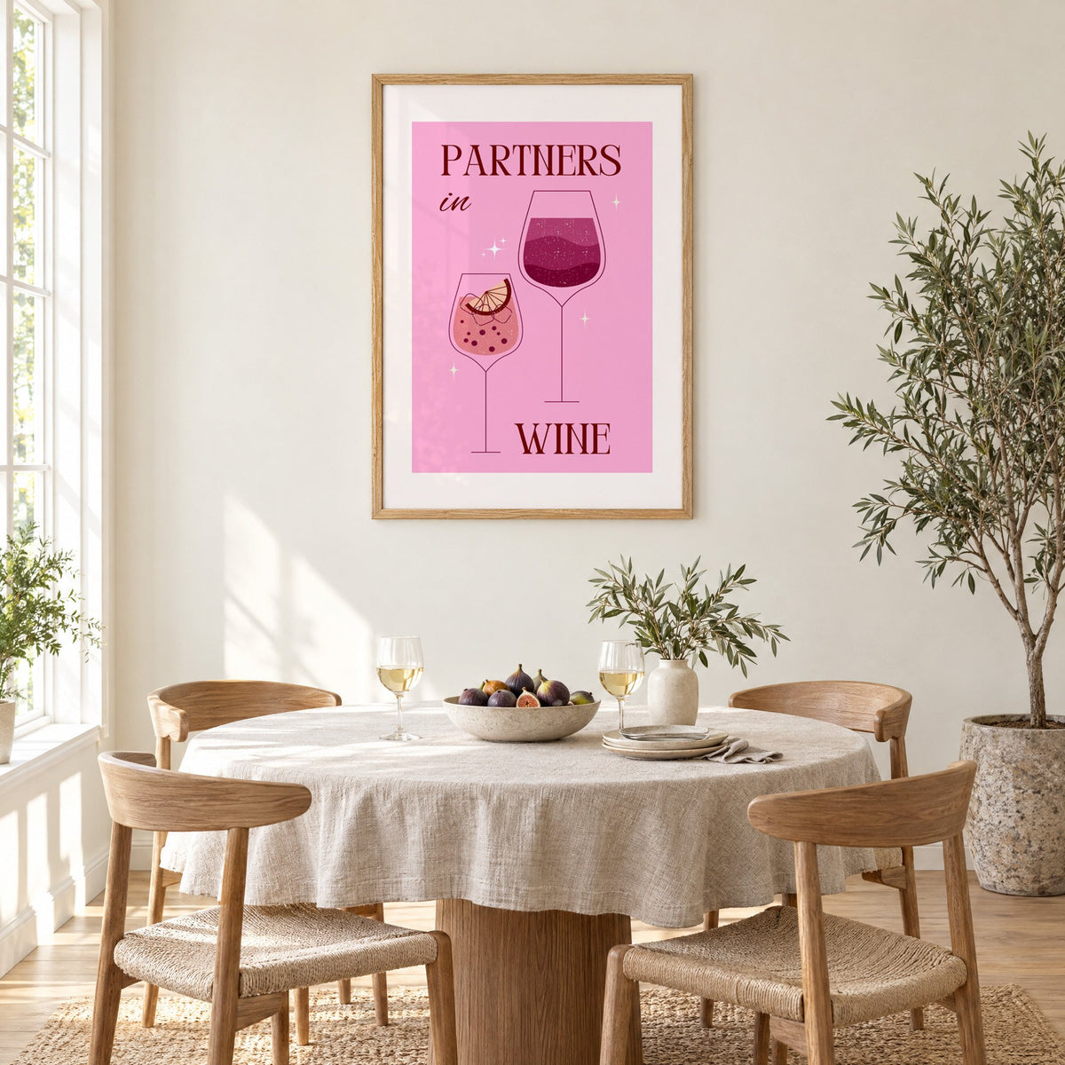 Partners in Wine two glasses pink print in an oak frame above a round linen-covered dining table with figs and wine
