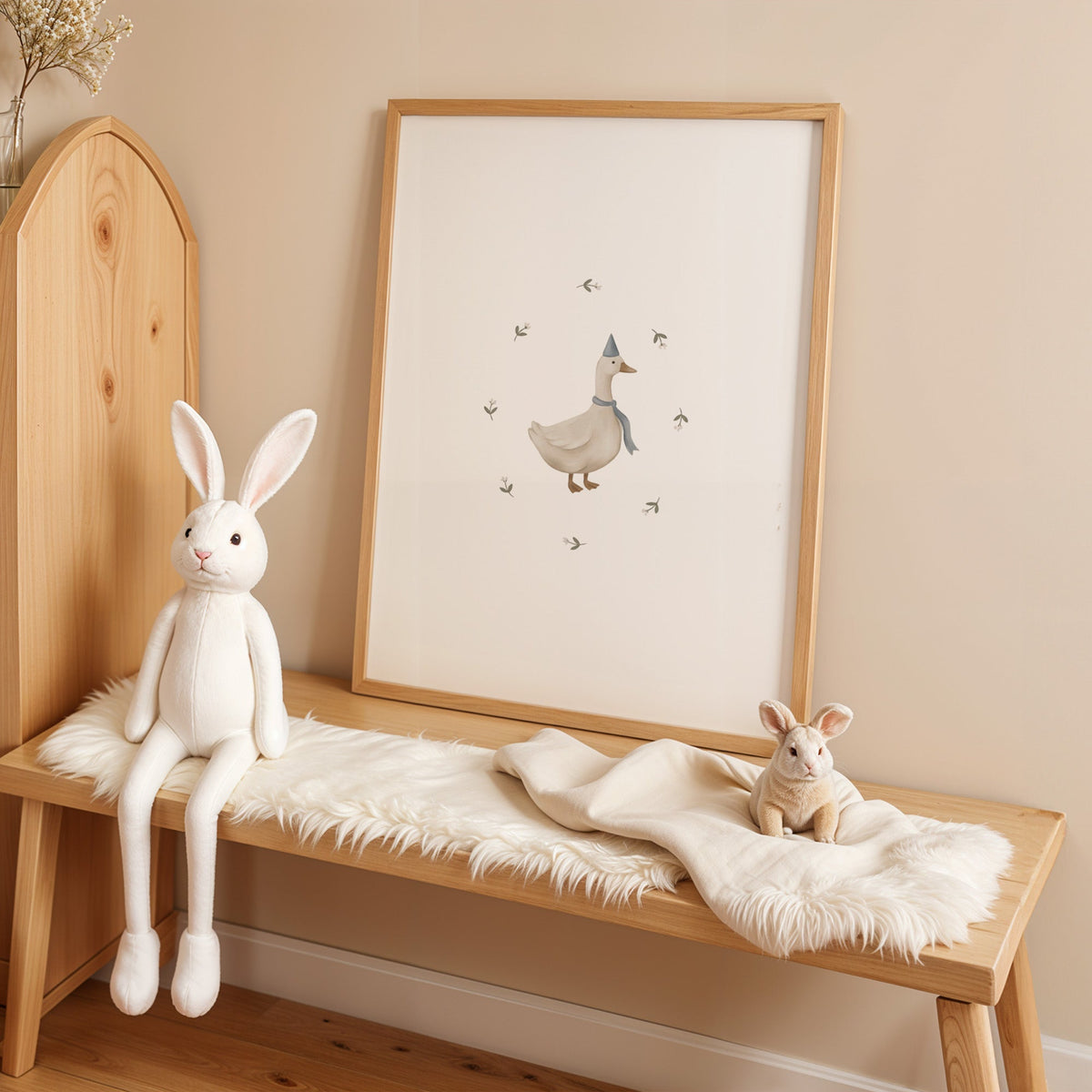 Goose in a blue party hat and scarf nursery print in an oak frame on a sheepskin bench beside a white plush bunny