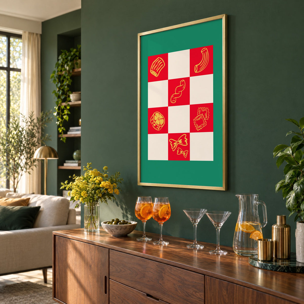 Red checkerboard pasta shapes print with green border in a brass frame on a bottle green wall above a sideboard with spritz and martini glasses, seen at an angle
