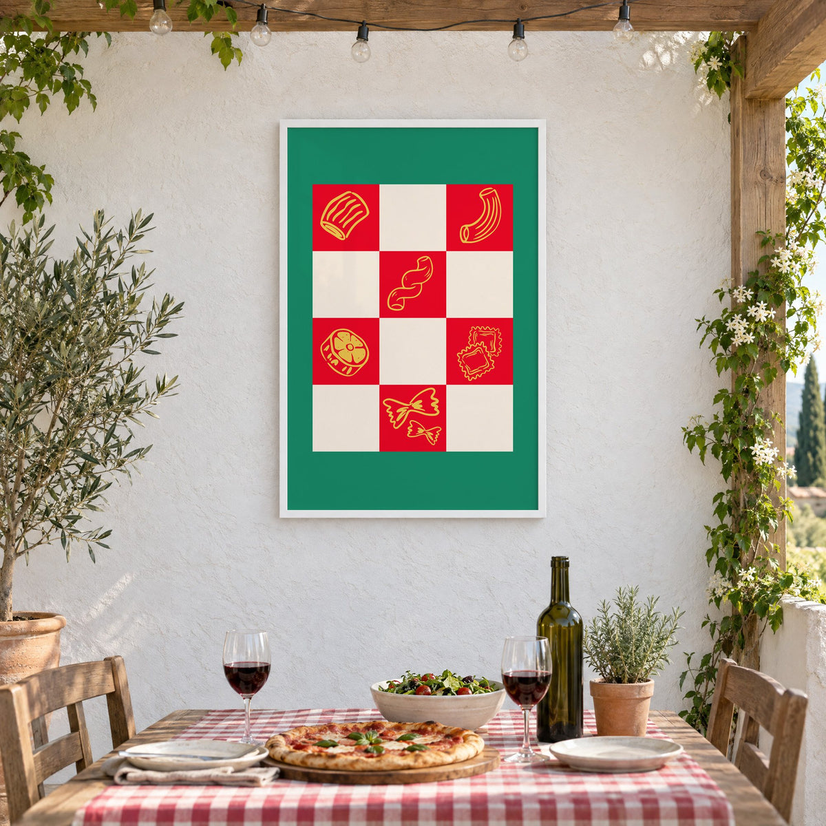 Red checkerboard pasta shapes print with green border in a white frame on a stucco wall above a checked-cloth table with pizza and red wine
