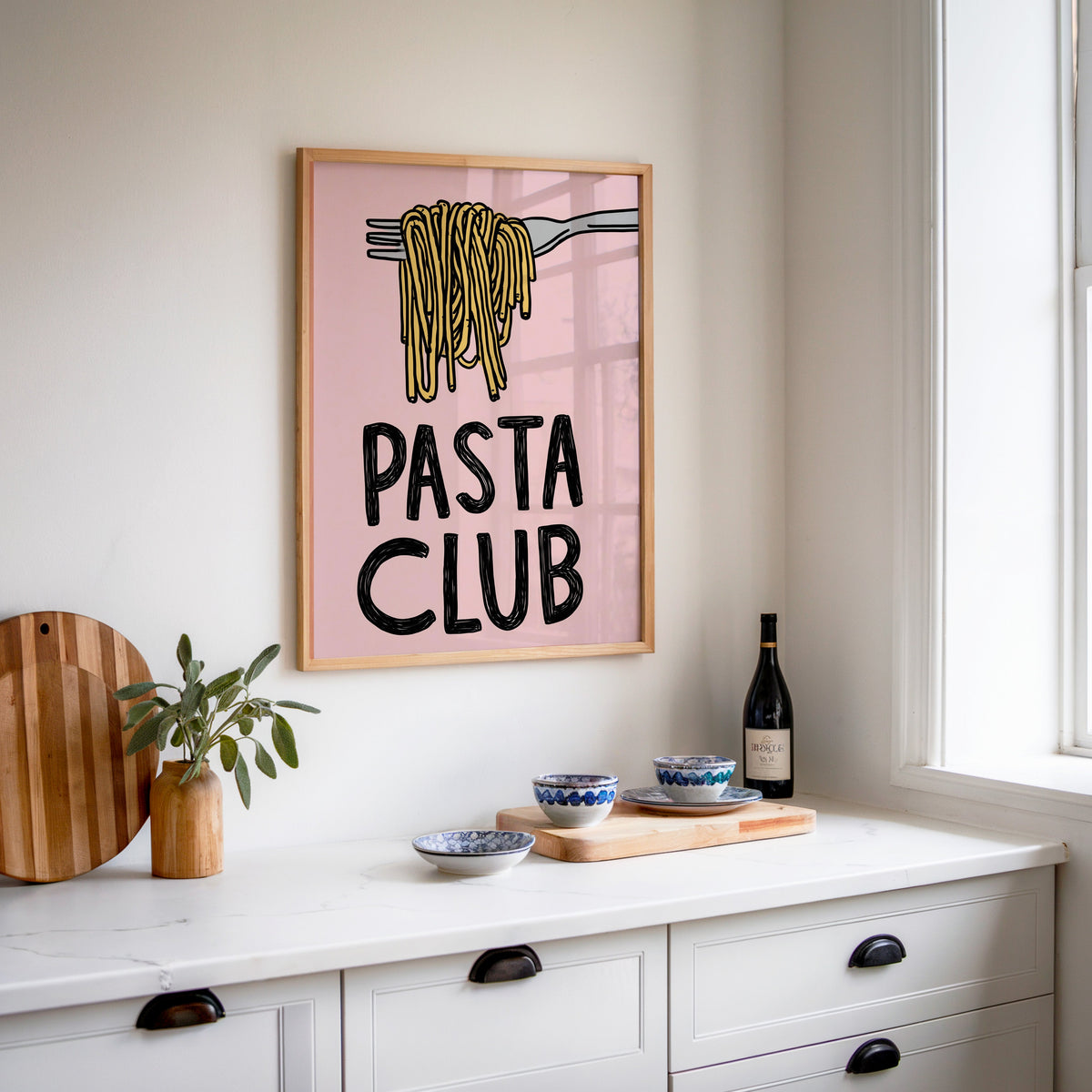 Pasta Club print in an oak frame above a white sideboard with a bottle of wine