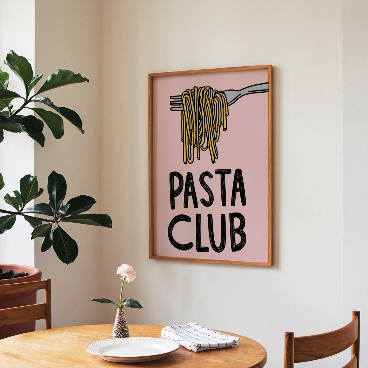 Pasta Club print in a walnut frame above a round table beside a fiddle-leaf fig