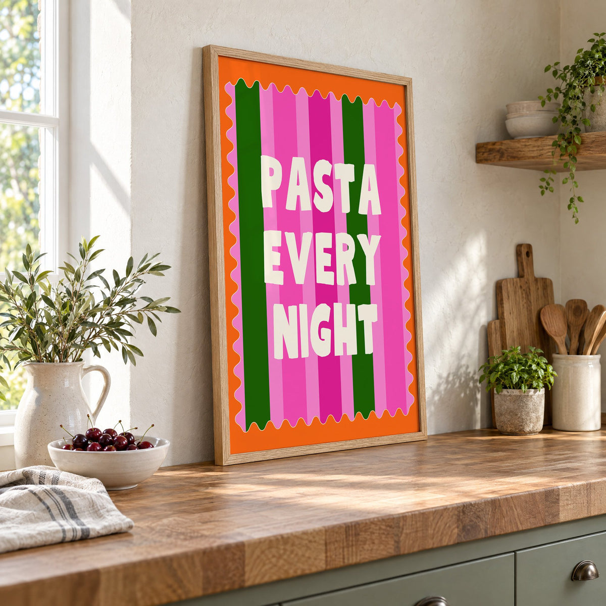 Pasta Every Night pink stripe print in an oak frame leaning on a butcher block island beside a bowl of cherries