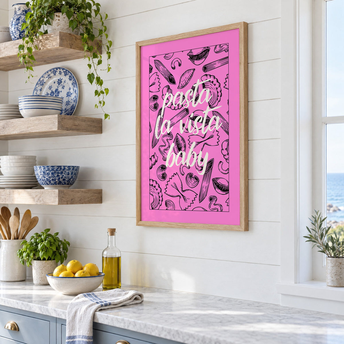 Pasta la vista baby pink print in an oak frame in a white coastal kitchen beside blue and white plates