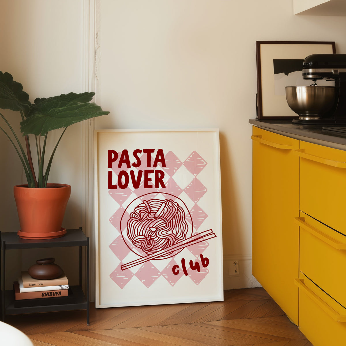 Pasta Lover Club print in a white frame leaning on parquet beside a bright yellow kitchen