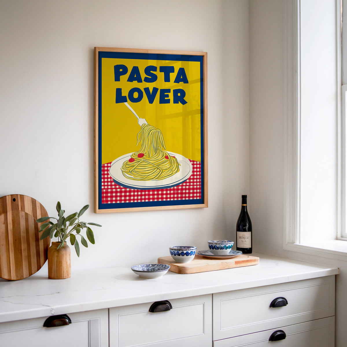 Pasta Lover yellow print in an oak frame above a white sideboard with a bottle of wine