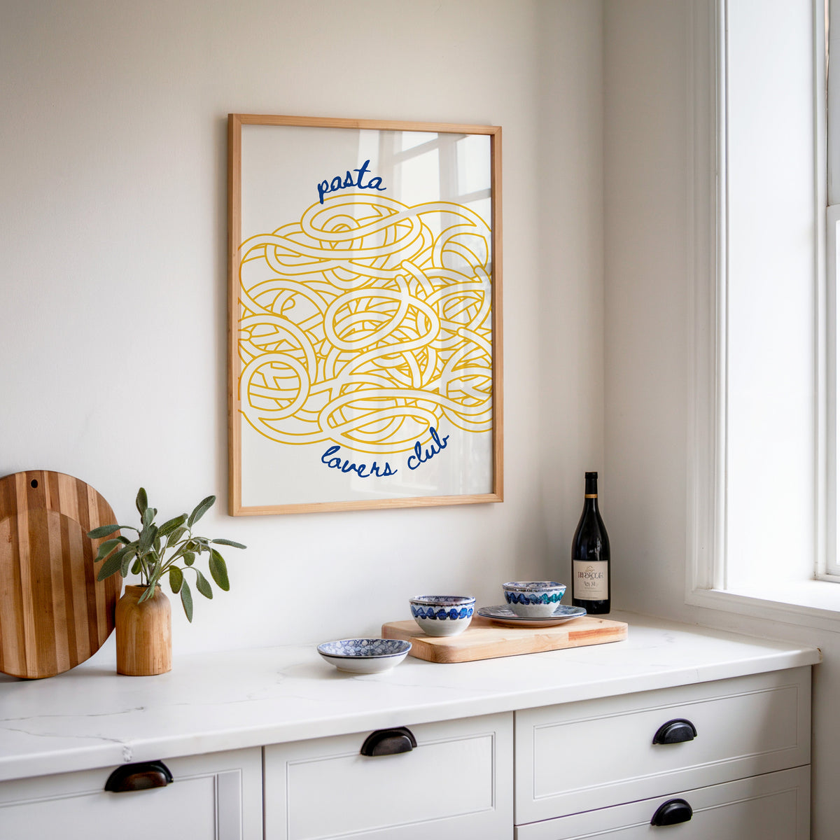 Pasta Lovers Club print in an oak frame above a white sideboard with a bottle of wine