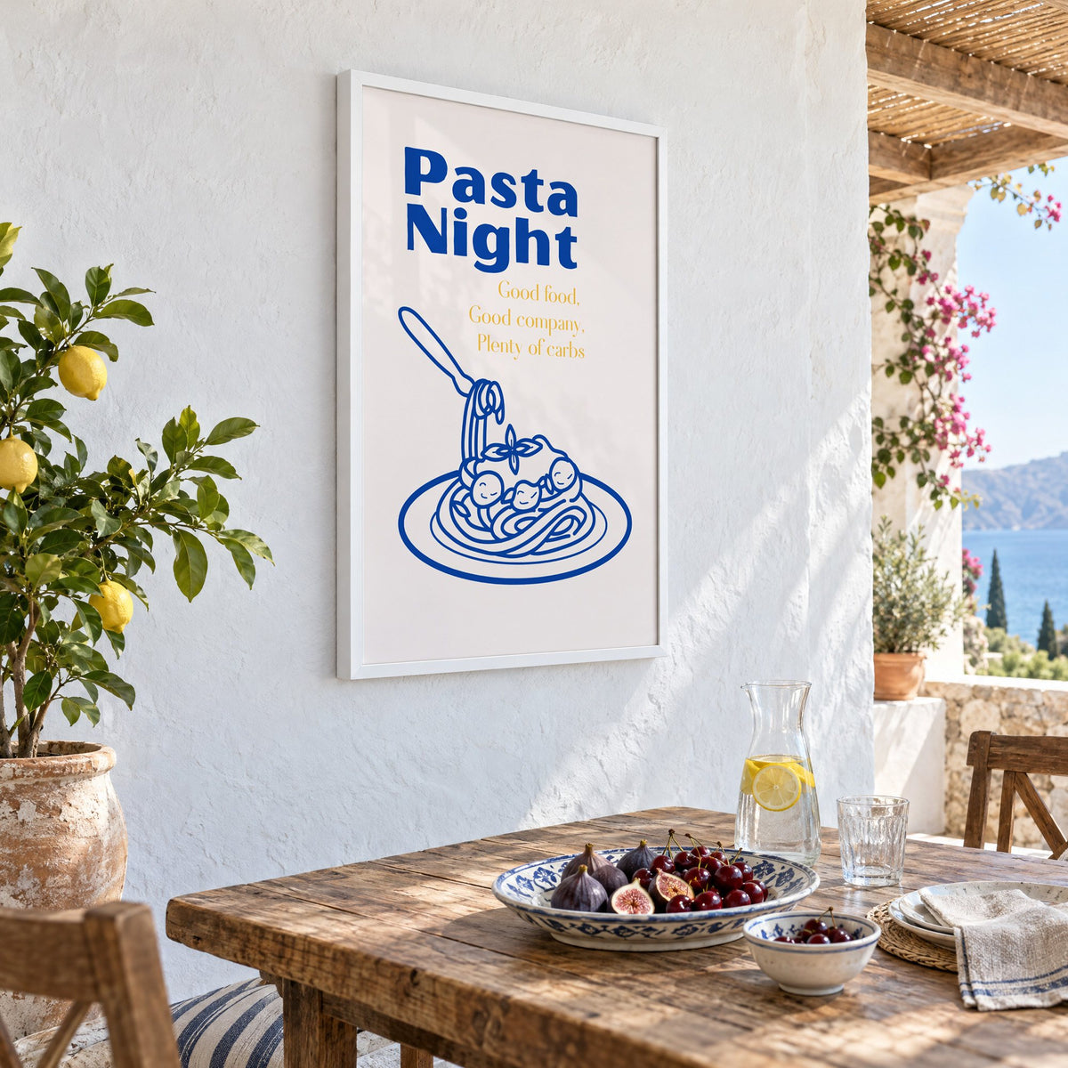 Pasta Night cobalt spaghetti and meatballs print in a white frame on a whitewashed terrace wall above a lunch table with figs and cherries, seen at an angle