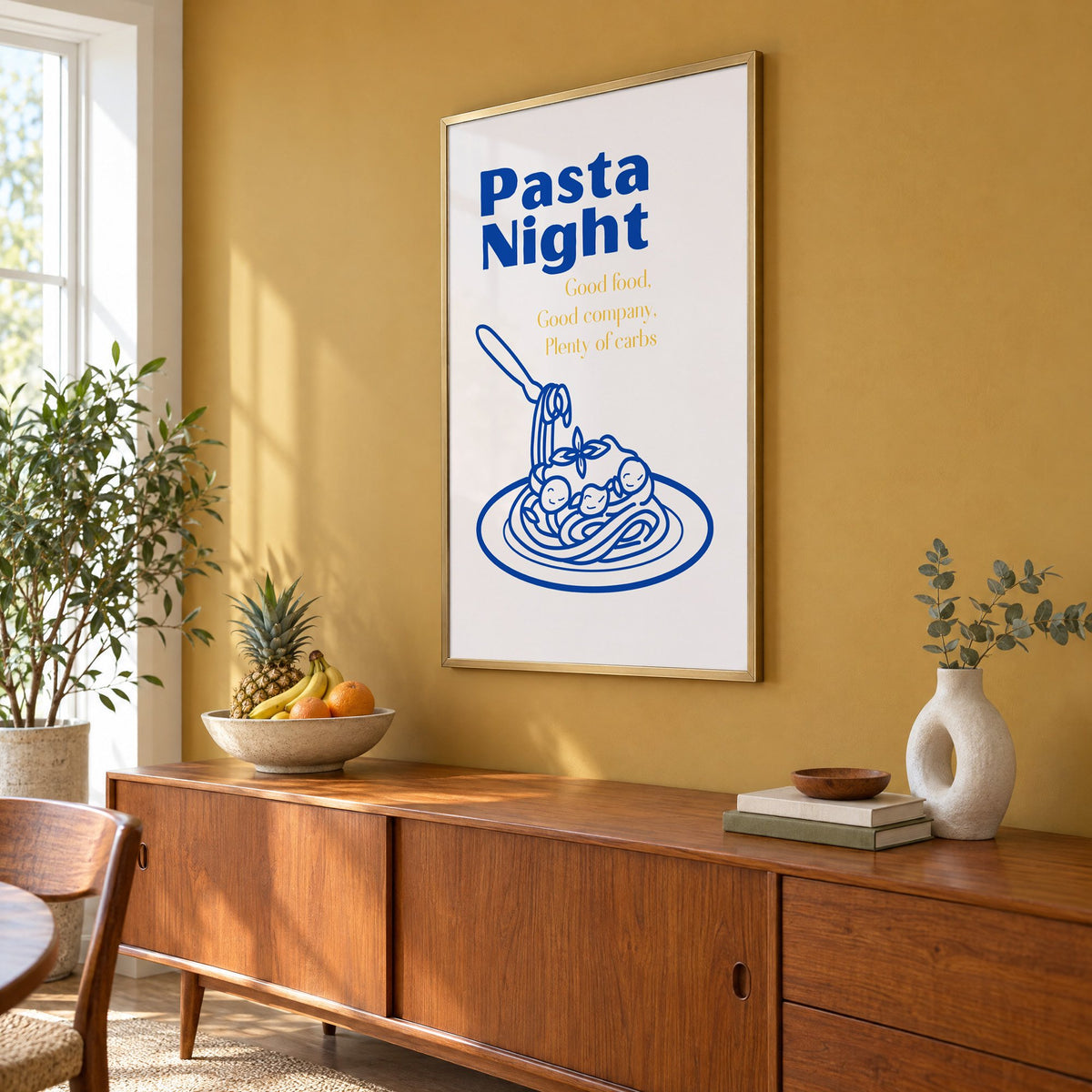 Pasta Night cobalt spaghetti and meatballs print in a brass frame on a mustard wall above a teak sideboard with a fruit bowl, seen at an angle