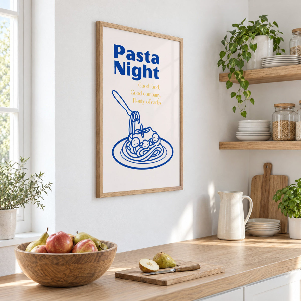 Pasta Night cobalt spaghetti and meatballs print in an oak frame on a white kitchen wall above a counter with apples and pears, seen at an angle