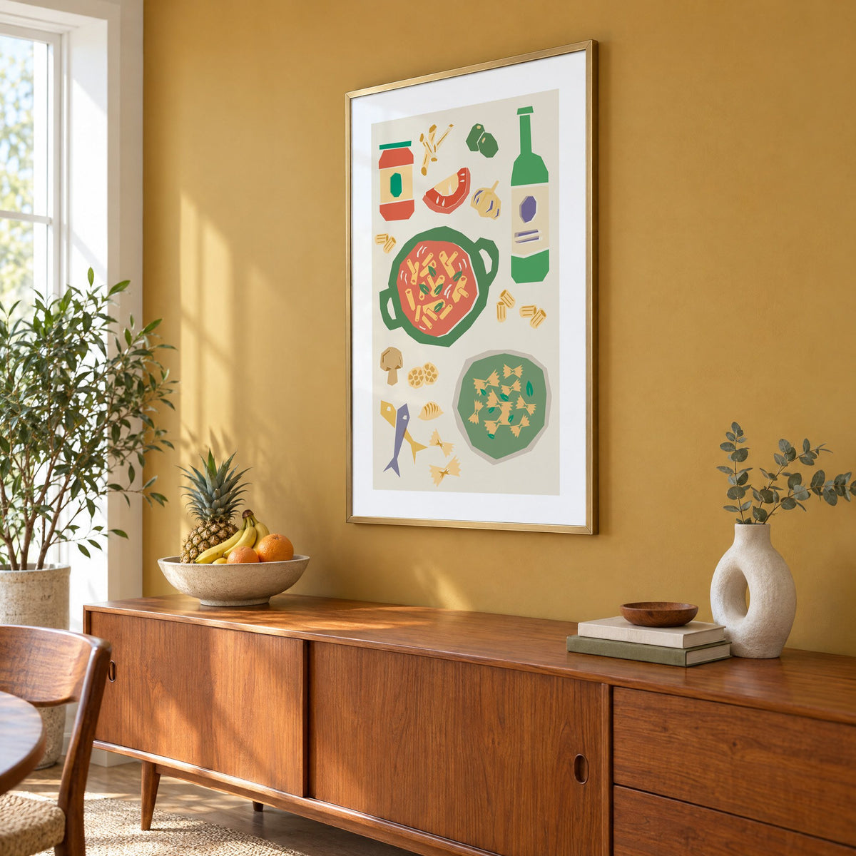 Penne pot and pesto farfalle cut-paper print in a brass frame on a mustard wall above a teak sideboard with a fruit bowl, seen at an angle