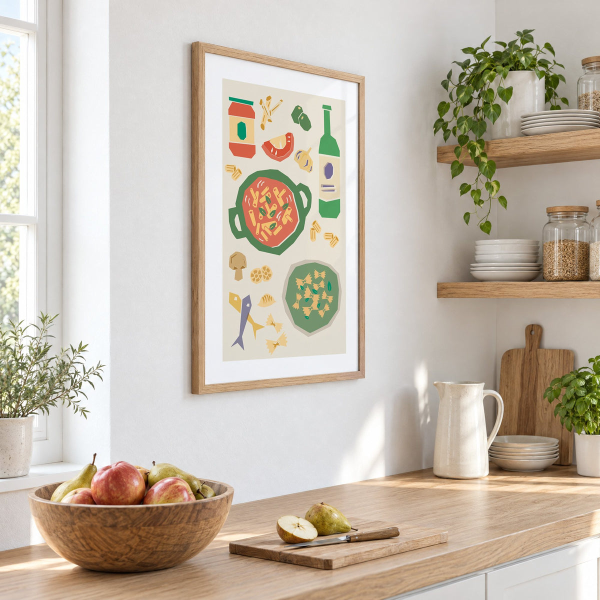 Penne pot and pesto farfalle cut-paper print in an oak frame on a white kitchen wall above a counter with apples and pears, seen at an angle