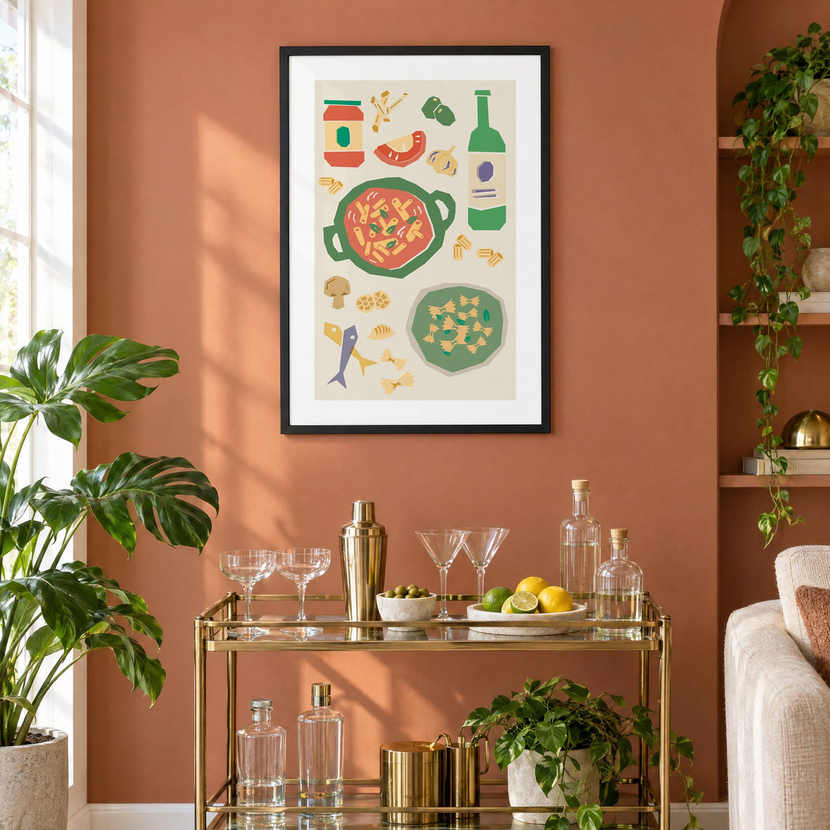 Penne pot and pesto farfalle cut-paper print in a black frame on a terracotta wall above a brass bar cart with coupes and citrus