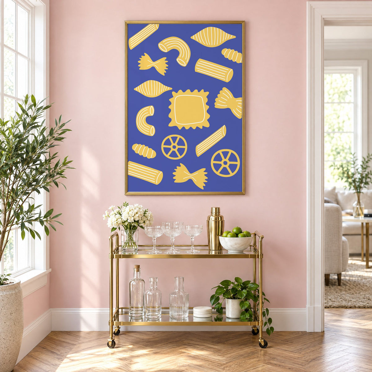 Yellow pasta shapes pattern on blue print in a brass frame on a blush pink wall above a gold bar cart with glassware