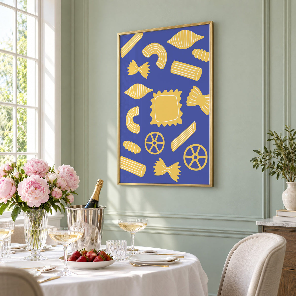 Yellow pasta shapes pattern on blue print in a brass frame on a sage wall above a brunch table with champagne and peonies, seen at an angle