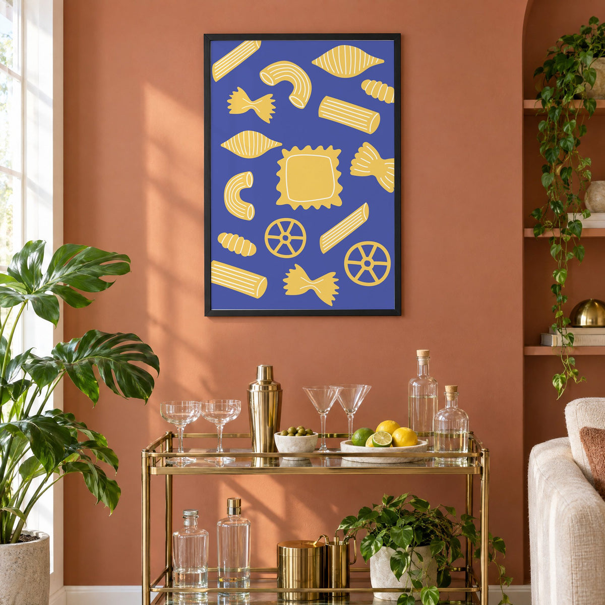 Yellow pasta shapes pattern on blue print in a black frame on a terracotta wall above a brass bar cart with coupes and citrus